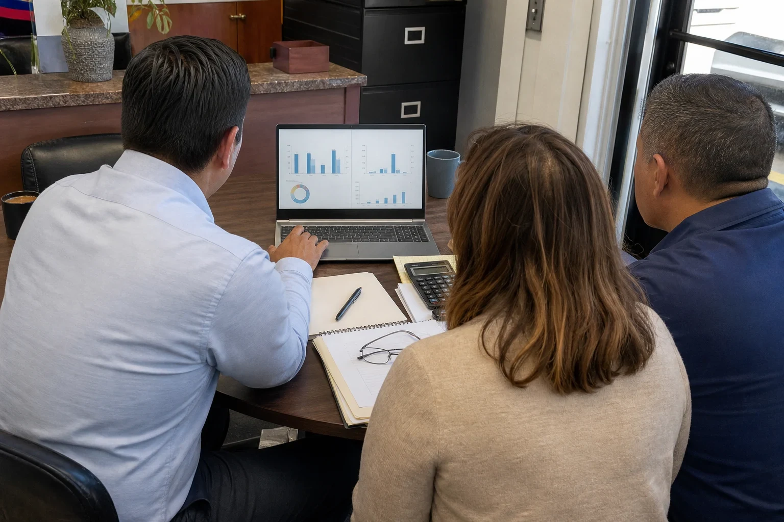 Agent in a light blue button-down shows charts on a laptop to a couple seated at a small round table in a storefront office beside a window and filing cabinet.