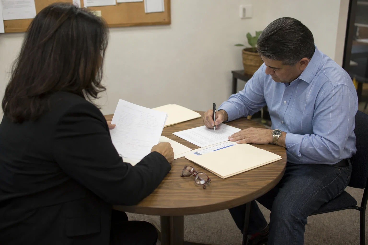 A man in a blue shirt signs a form at a round wooden table while a woman in a black blazer reviews papers, with manila folders and reading glasses on the table