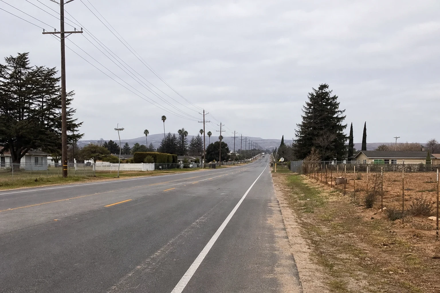 Quiet two-lane road on the edge of town under an overcast sky, with power lines, palm and pine trees, single-story homes, a fenced dirt lot and hills beyond